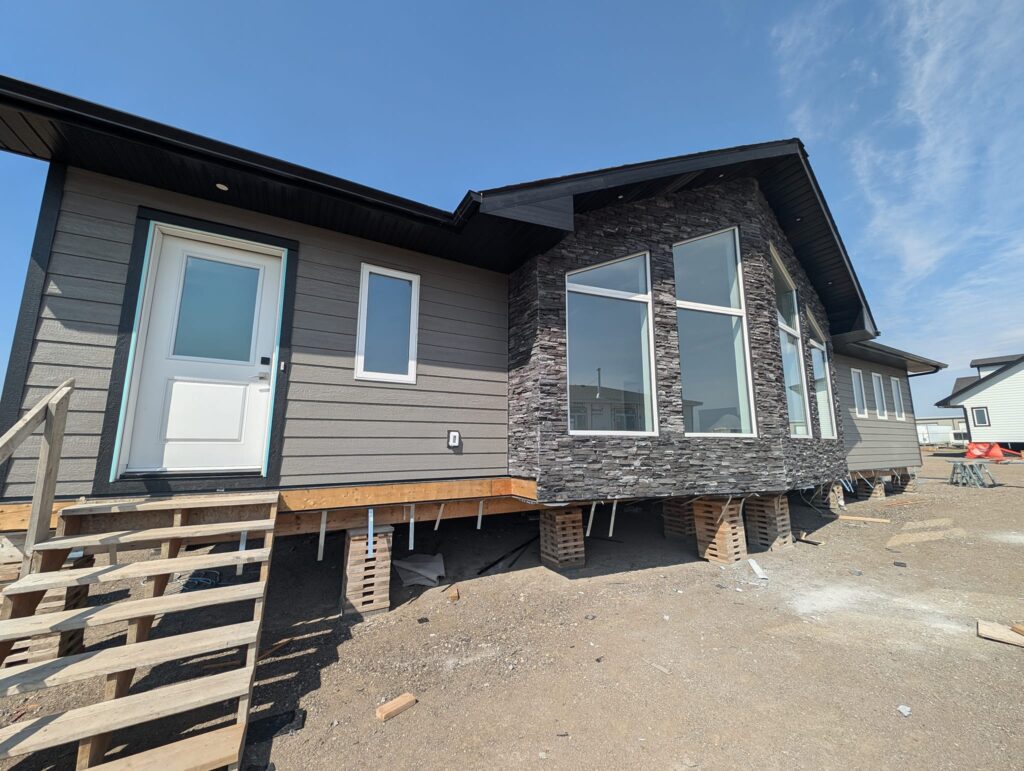 Modern modular home under construction with gray siding and stone accents. Large windows and temporary wooden steps create a sense of potential and progress.