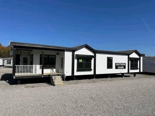 A single-story white modular home with dark trim, labeled "Southern Charm," set on a gravel lot under a clear blue sky, exuding a rustic, inviting feel.