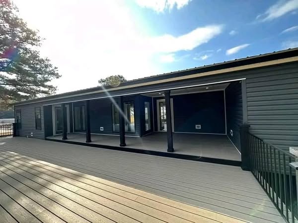 Wide wooden deck with a modern dark blue house featuring large windows and a covered porch. Sunlight casts soft flares, creating a tranquil atmosphere.