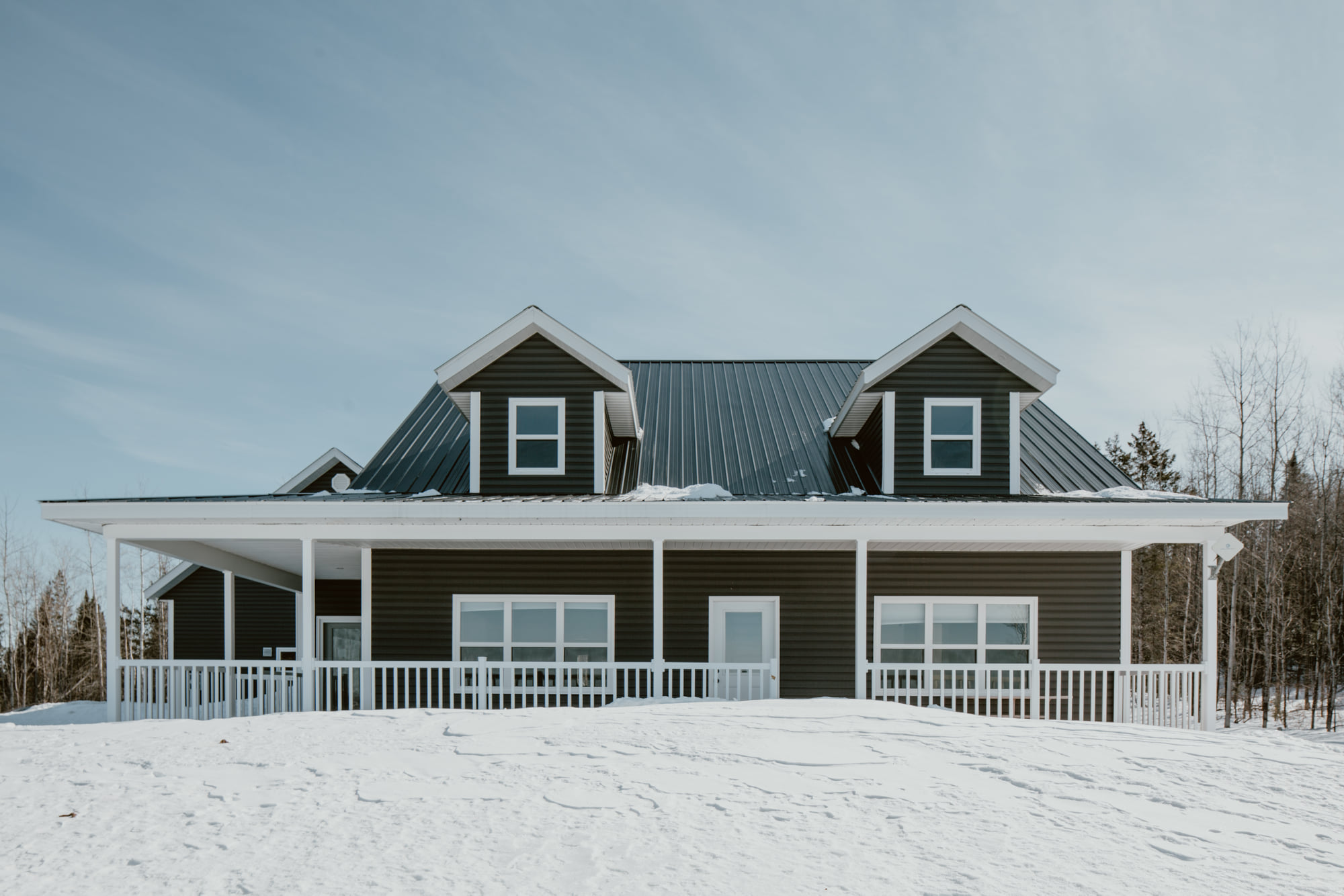 A dark green house with a steep metal roof and white trim sits in a snowy landscape. The wraparound porch and clear sky create a serene, wintery feel.