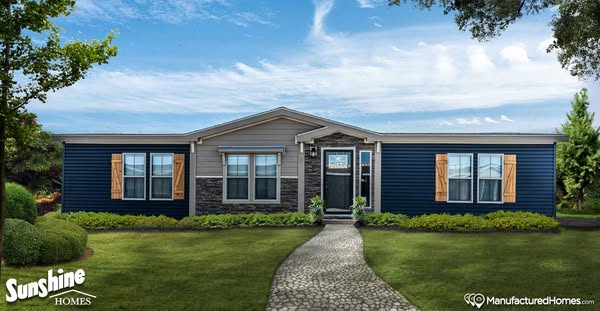 A modern manufactured home with blue and gray siding, wooden shutters, and stone accents. A cobblestone path leads through a lush, green lawn.