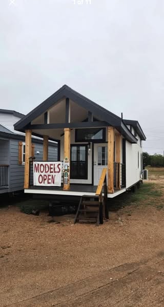 Small modern tiny house with a gabled roof and wooden accents. A "Models Open" sign hangs on the porch, set against a cloudy sky in a dirt lot.