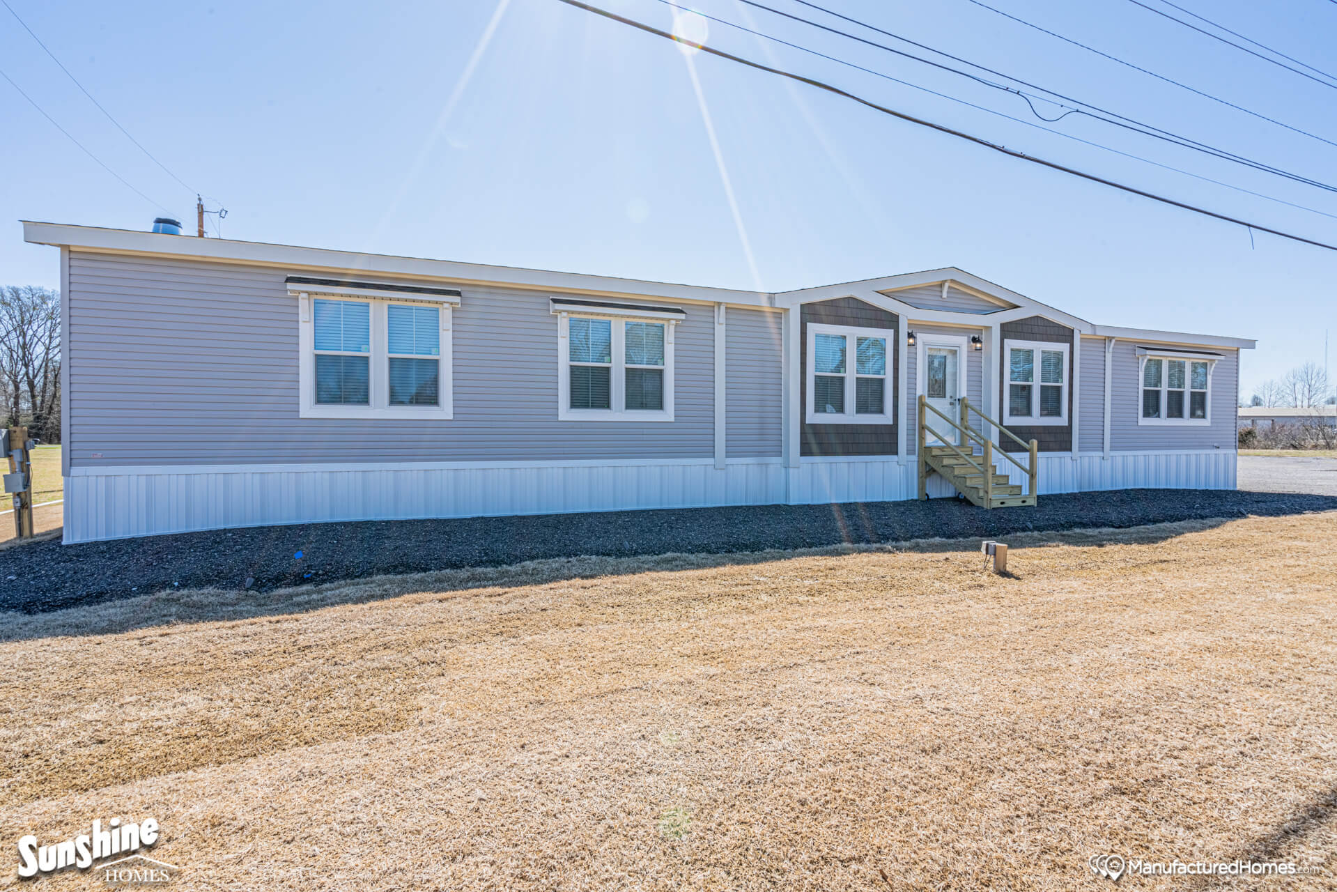 A modern, single-story manufactured home with beige siding and a small porch, set on a sunny day with clear skies and dry grass in the foreground.