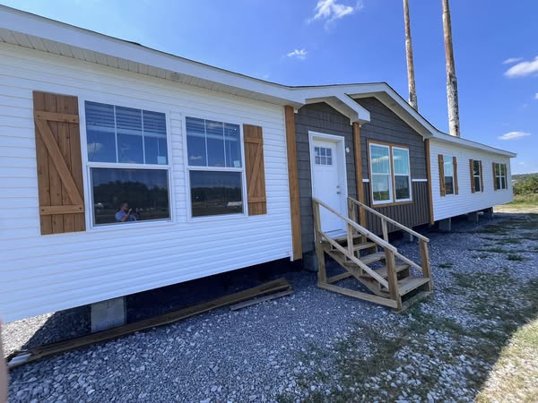 A white and brown modular home with wooden shutters stands on a gravel base under a clear blue sky. Wooden steps lead to the front door.