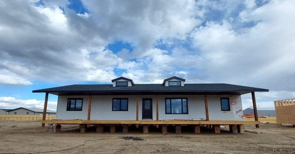 A single-story house on stilts under a partly cloudy sky. The house is unfinished, with a wide porch and exposed beams, conveying a sense of progress.