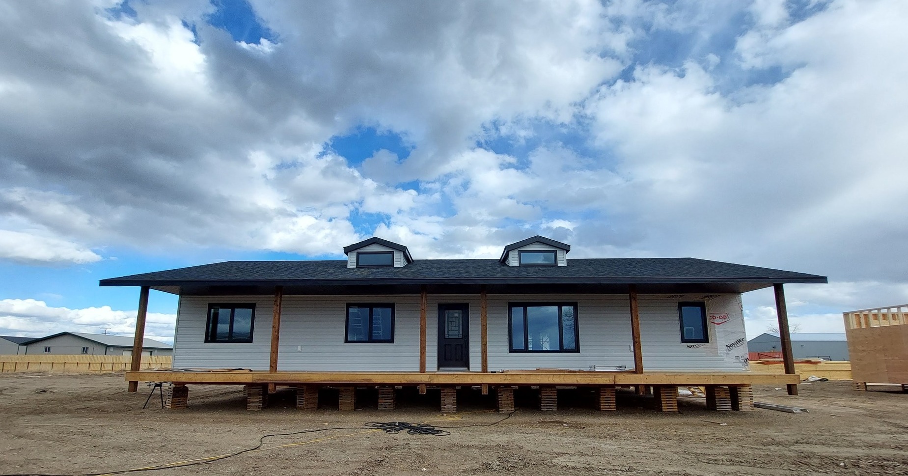 A single-story house on stilts under a partly cloudy sky. The house is unfinished, with a wide porch and exposed beams, conveying a sense of progress.