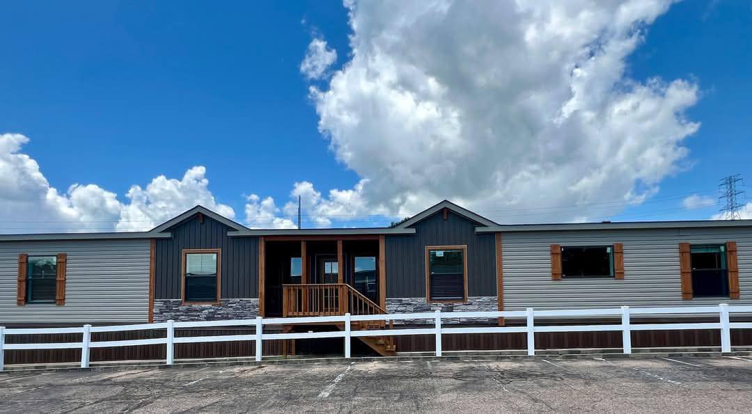 A modern modular home with a gray exterior, wooden accents, and a front porch. It is set against a bright blue sky with scattered clouds, bordered by a white fence.