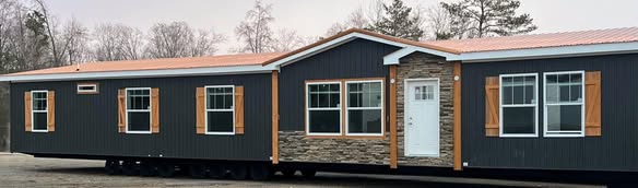 A dark gray mobile home with wooden shutters and a stone accent around the entrance, set in a wintry landscape with leafless trees in the background.