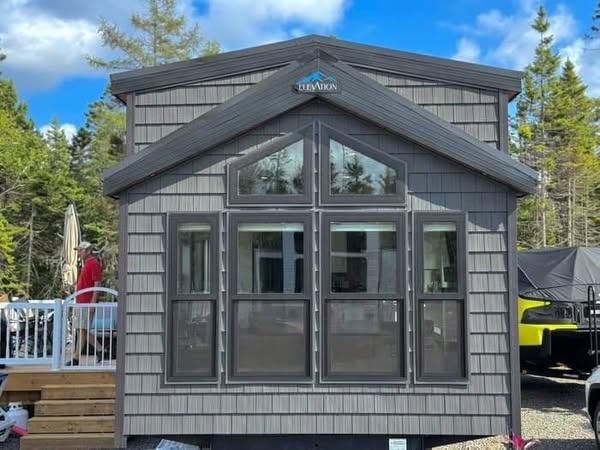 Cozy tiny house with gray siding and large front windows, nestled among lush green trees under a bright blue sky. Peaceful and inviting atmosphere.