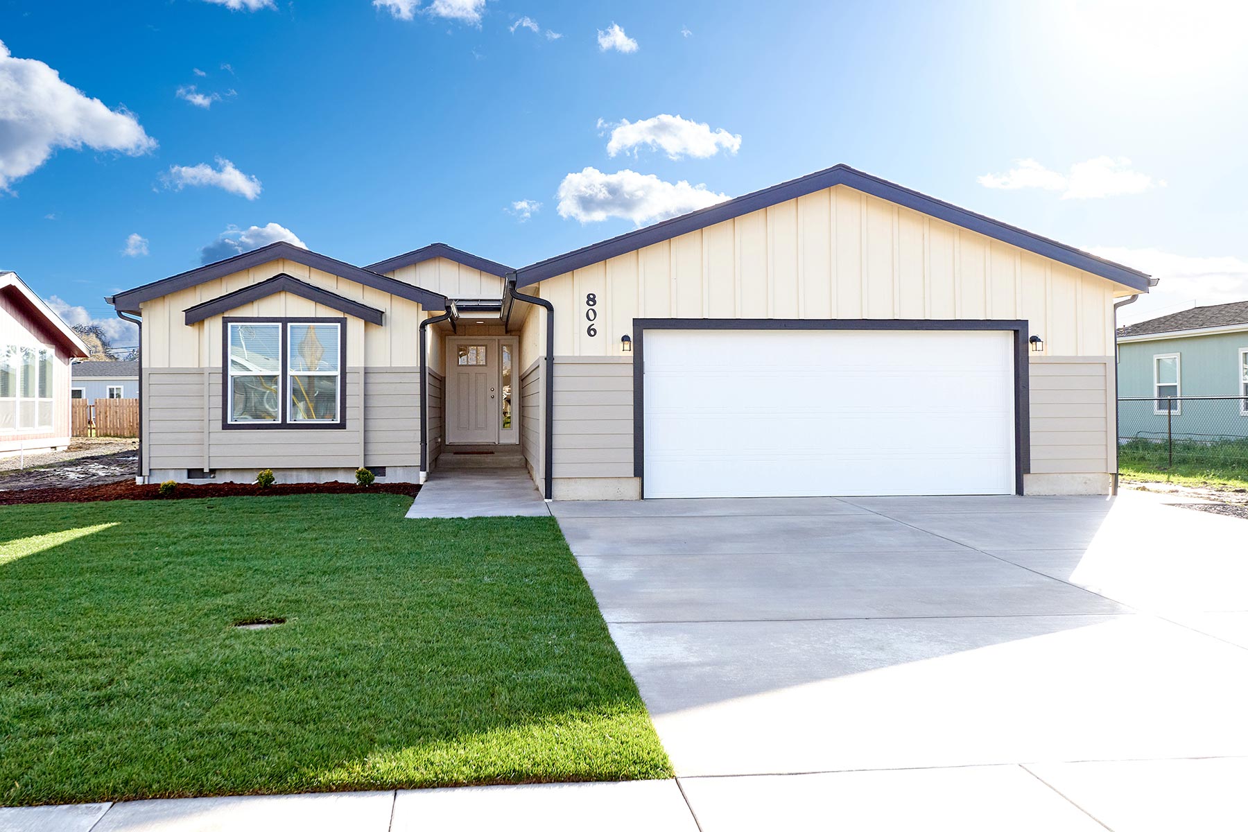 A modern, single-story house with beige siding and a white garage is fronted by a neat lawn under a bright blue sky with fluffy clouds. The atmosphere is serene.