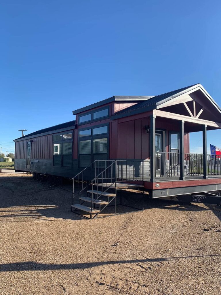 A modern tiny house with red and gray siding sits on a gravel lot under a clear blue sky. It features large windows, a metal roof, and a small porch with steps.