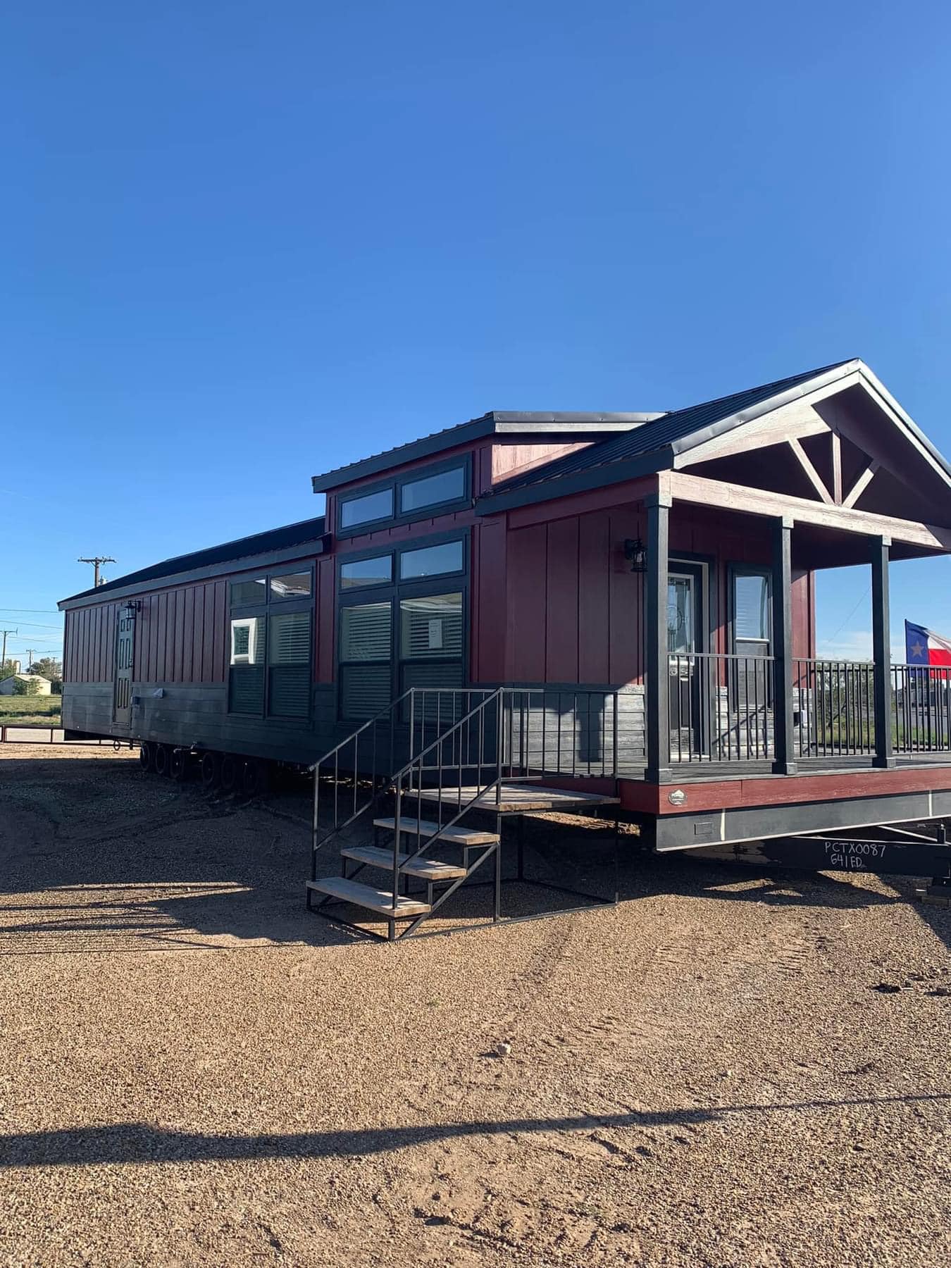 A modern tiny house with red and gray siding sits on a gravel lot under a clear blue sky. It features large windows, a metal roof, and a small porch with steps.
