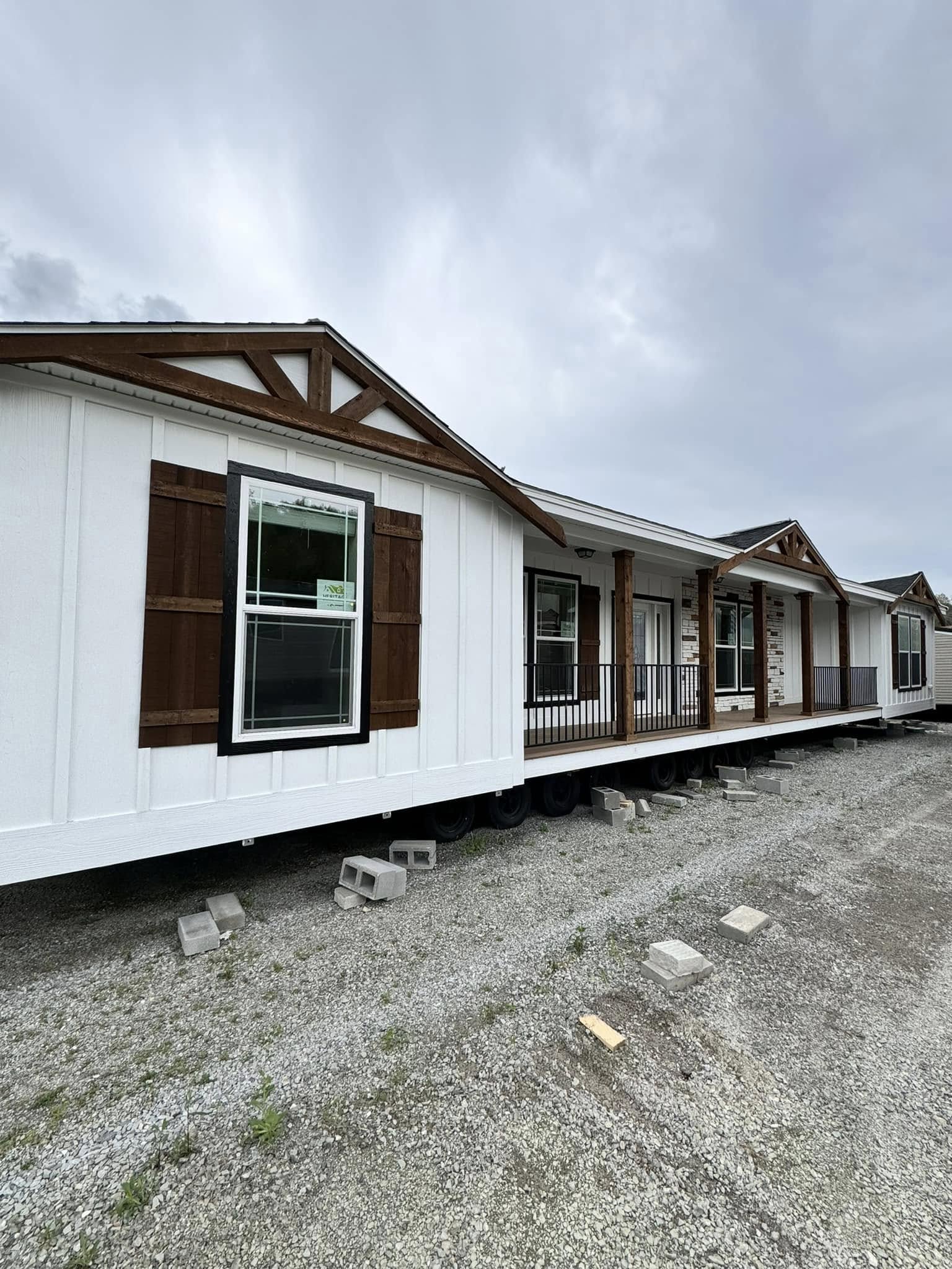 Alt text: "A prefabricated house featuring a white exterior with wooden accents and a small porch is set on concrete blocks against a cloudy sky."