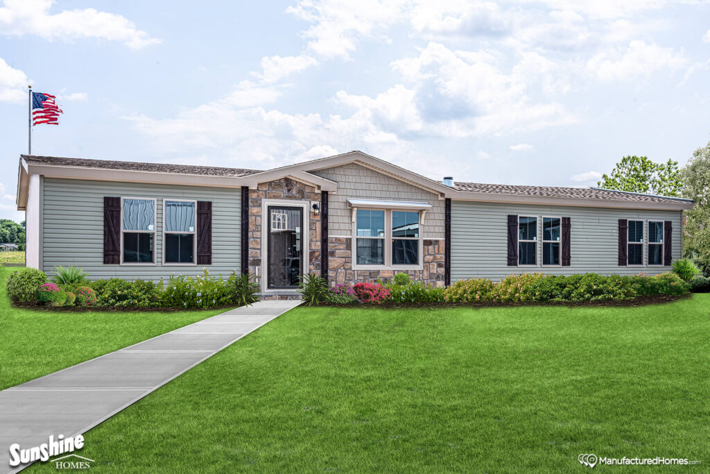 A single-story manufactured home with gray siding and stone accents stands amidst a lush green lawn. An American flag waves on the left, conveying a welcoming and patriotic tone.