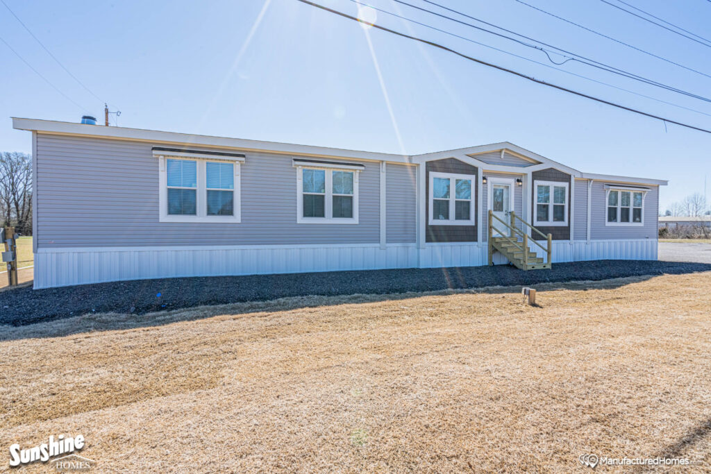 Single-story manufactured home with gray siding and white trim, set against a clear blue sky. Wooden steps lead to the front door, situated in a grassy area.