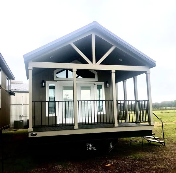 Small modern tiny house with white-trimmed porch and black railing, set on a grassy field. Overcast sky enhances a cozy, inviting atmosphere.