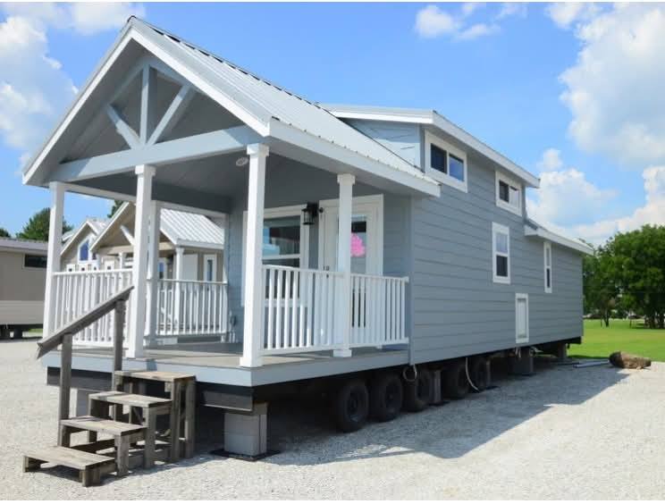 A small gray mobile home with a white porch and railing sits on wheels. It has a metal roof and a few windows. The sky is clear and sunny.