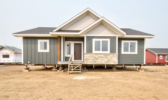 Single-story house under construction on a dirt lot, featuring gray siding, white-framed windows, and a small wooden porch, conveying a sense of progress.