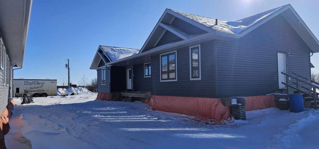 Single-story blue house with snow-covered roof, surrounded by snow and a clear blue sky. An orange tarp is draped around the base, and a white trailer is visible nearby.