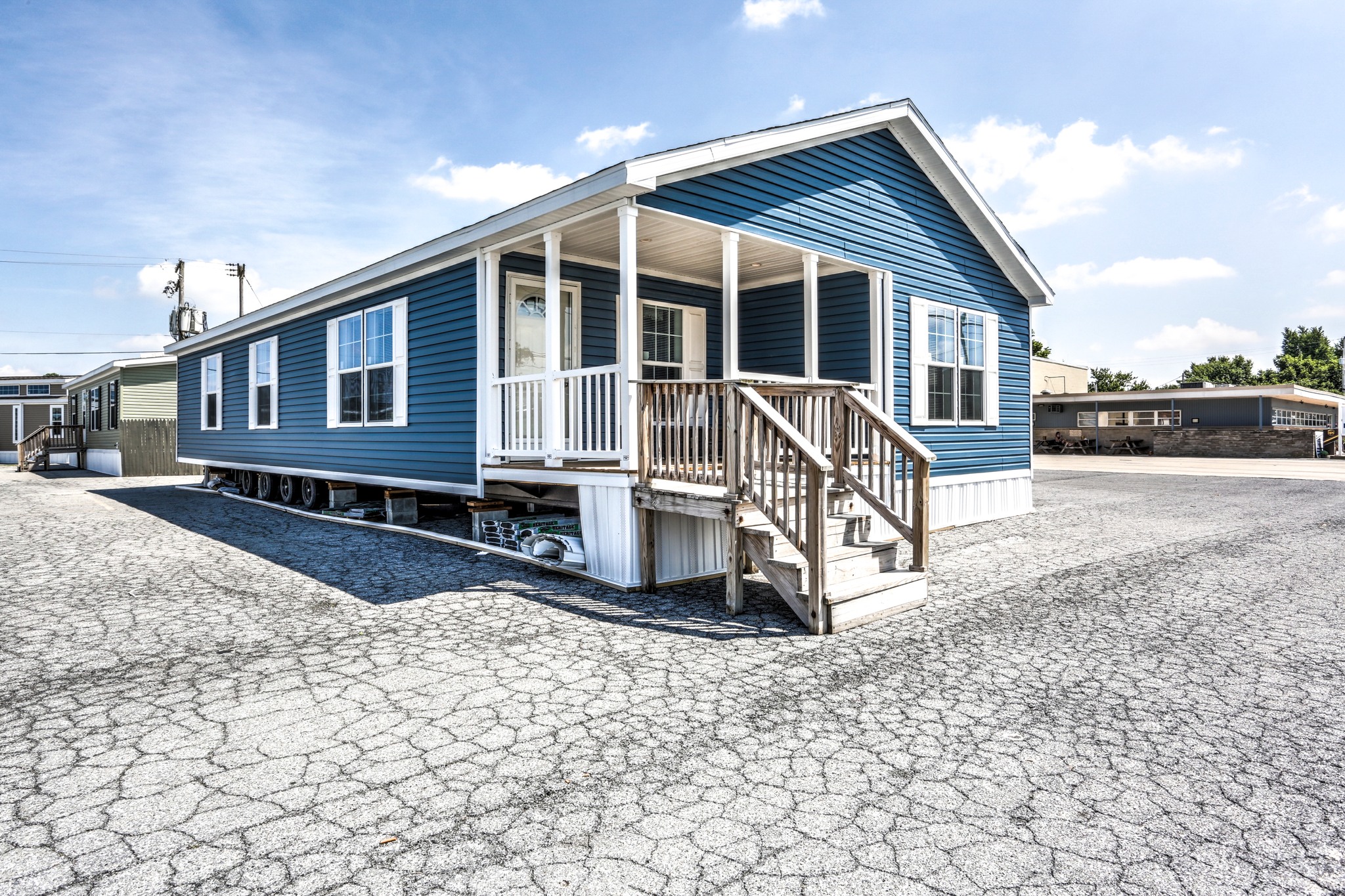 Blue manufactured home with a small covered porch and stairs, set on a gravel lot under a sunny sky. The scene conveys a sense of simplicity and openness.