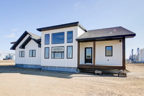 Modern, single-story, white modular house with dark trim, large windows, and a small porch, set in a sunny, spacious, semi-rural environment.