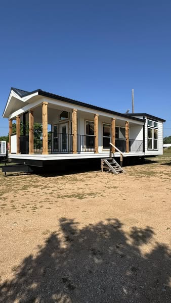 Modern tiny home with a sloped roof, wooden pillars, and large windows under a clear blue sky. A small set of stairs leads to the entrance.