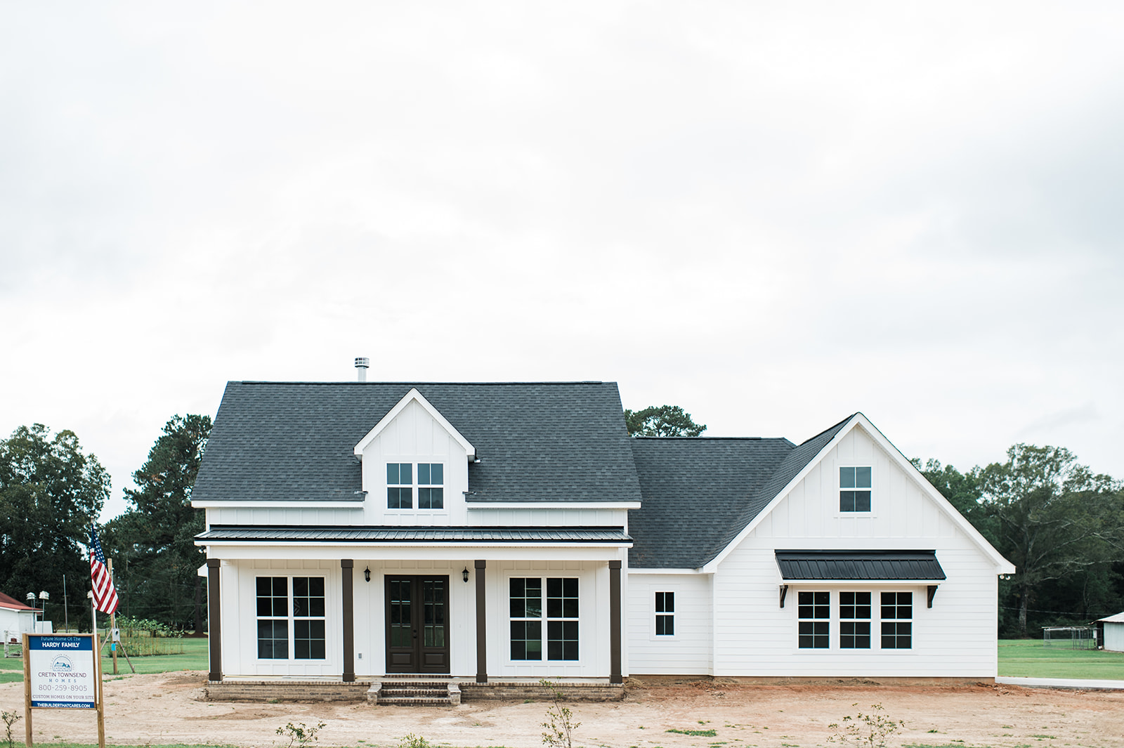A white farmhouse-style house with a dark roof sits on a dirt lot. It features a front porch, new windows, and a neatly trimmed lawn, evoking a sense of simplicity and charm.