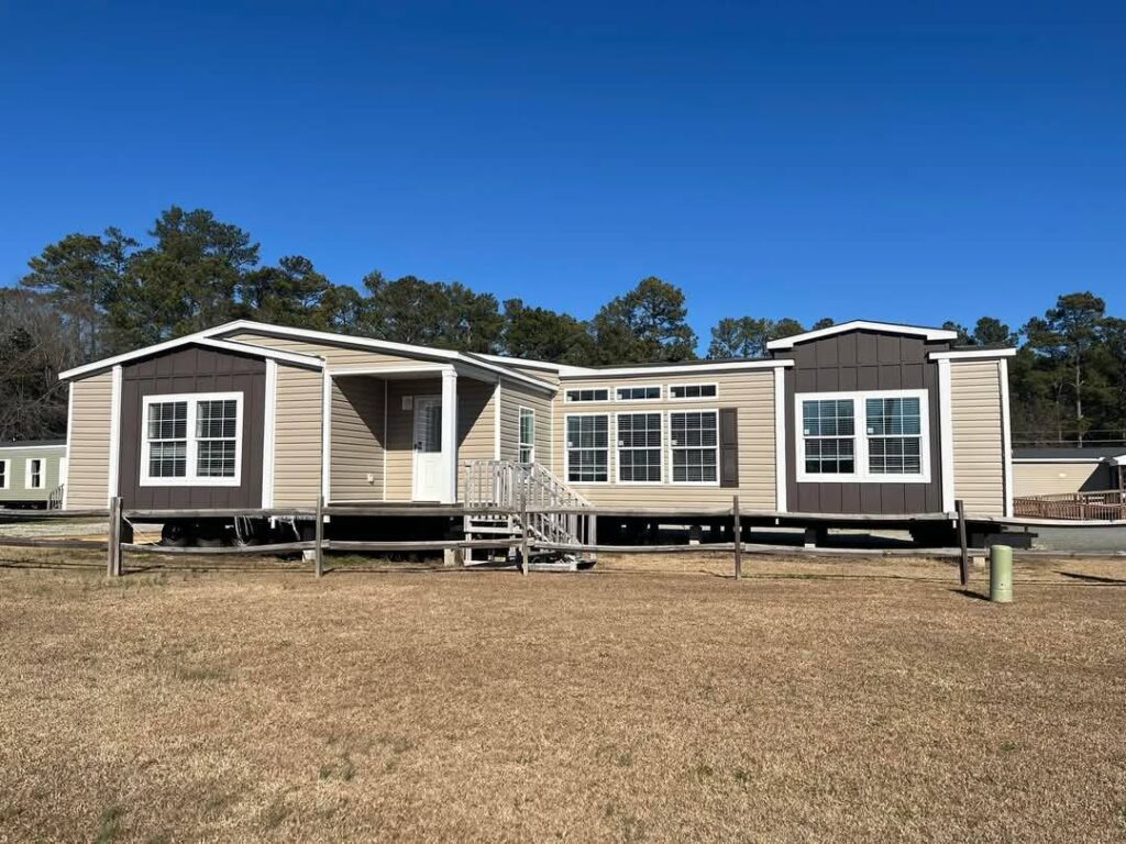 A beige double-wide modular home with brown accents sits on a grassy lot. Large windows, small stairs, and clear sky in the background convey a serene, open atmosphere.