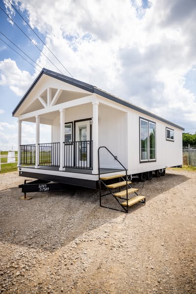 Small white mobile home with a front porch, black railing, and steps. It's positioned on a gravel lot under a partly cloudy blue sky.