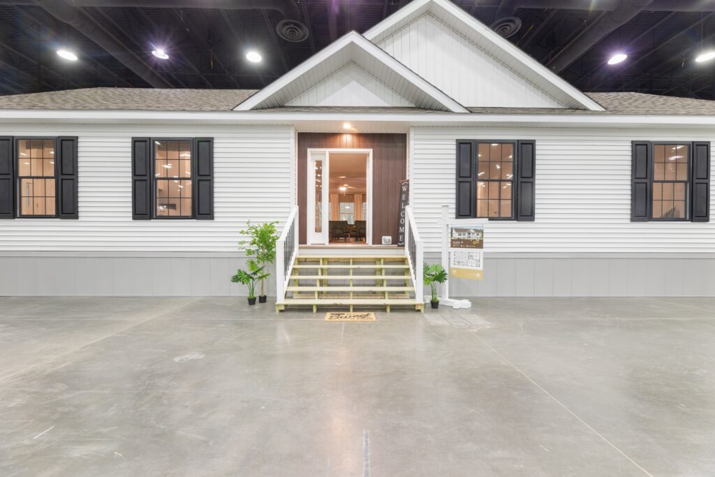Front view of a modern white show home inside a building, featuring gray trim, black shutters, a gabled roof, steps, and potted plants.