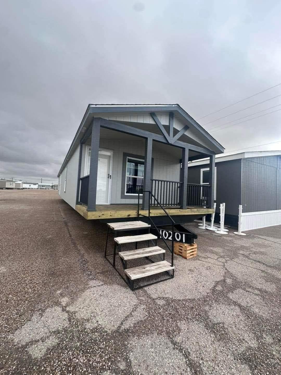 A modern gray mobile home with a small porch and railing, positioned on a gravel lot under an overcast sky, conveying a minimalist, calm atmosphere.