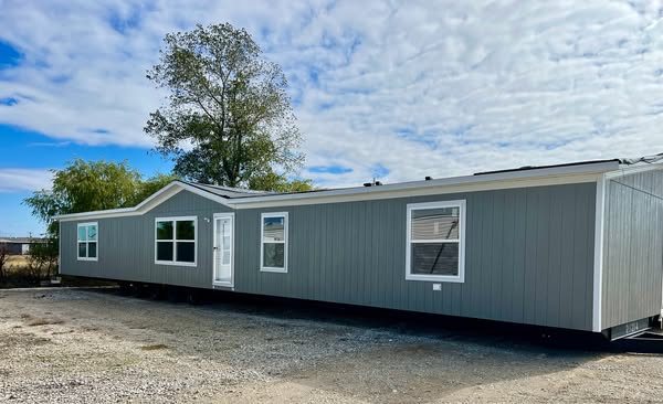 A gray mobile home with large windows and a white door sits on gravel, set against a partly cloudy sky. Trees surround it, creating a serene atmosphere.