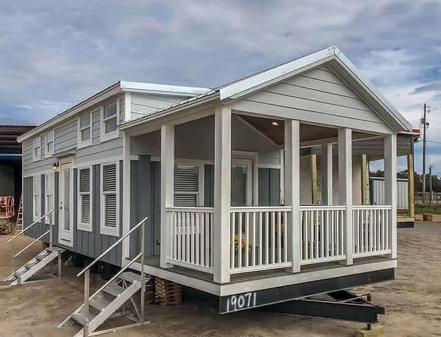 A small, grey and white modular home with a gabled roof and a covered porch. Two sets of steps lead to the porch. The sky is overcast, suggesting a calm setting.
