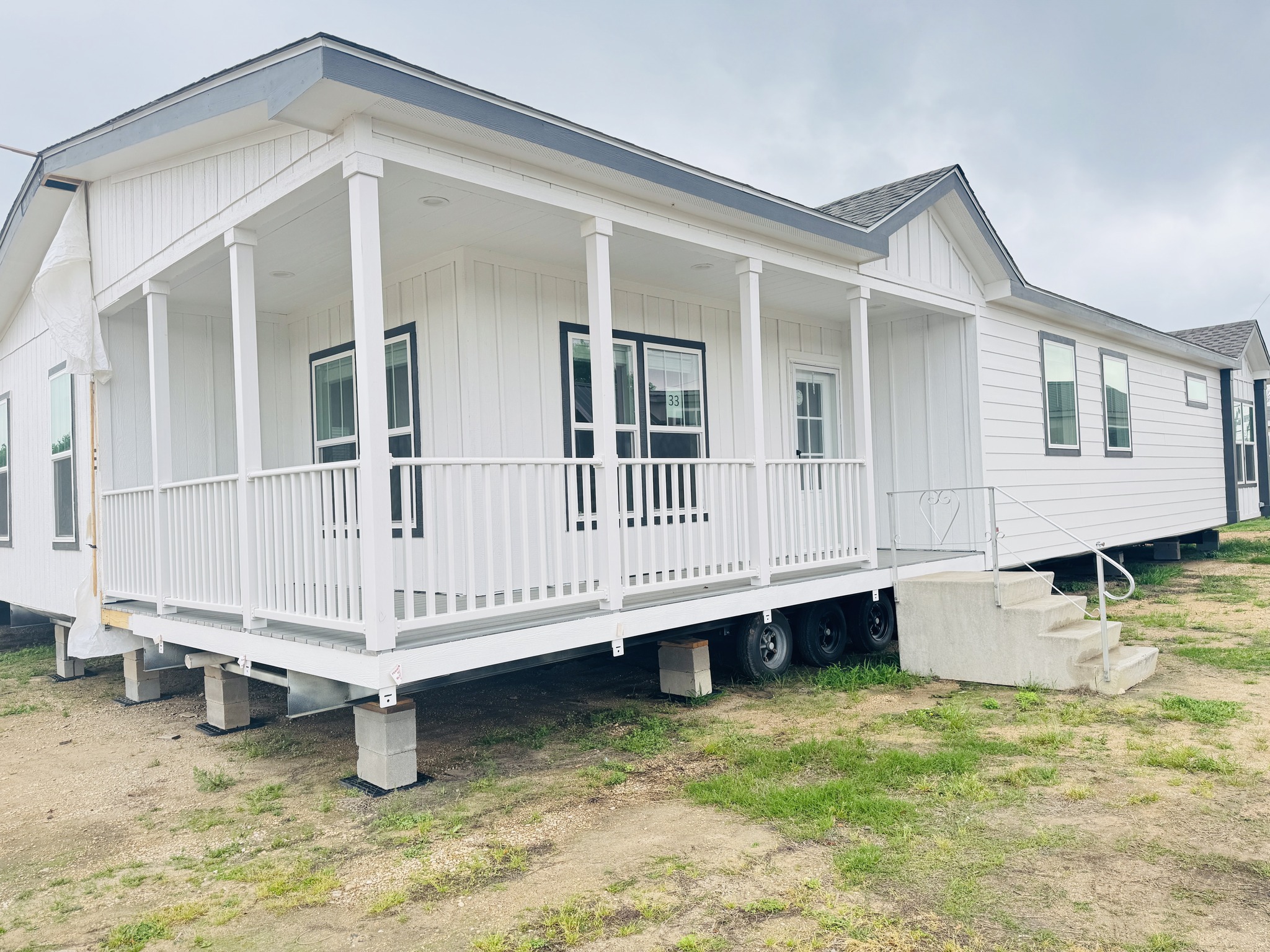 A new white manufactured home with a covered porch, black-trimmed windows, and steps leading up. It sits on grass under a cloudy sky, conveying a fresh and inviting setting.