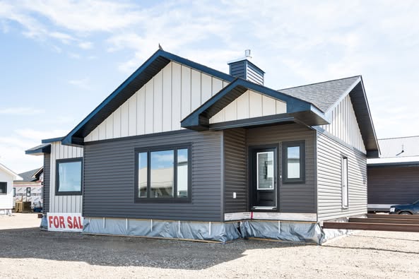 Single-story house with dark siding and pitched roof against a blue sky. Large windows and a "For Sale" sign add a feeling of opportunity.