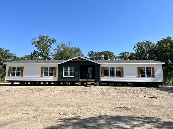 Single-story modular home on dirt lot with white siding, green central section, wooden shutters, and surrounded by trees under clear blue sky.