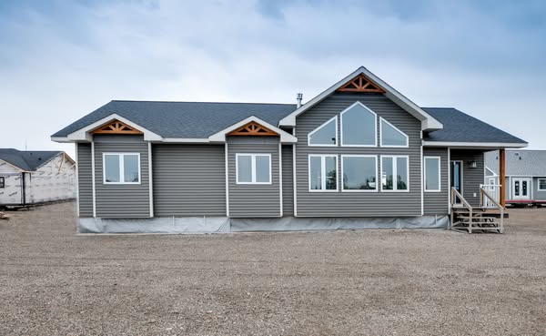 A gray, modern modular home with large windows and a gabled roof stands on a gravel lot under a cloudy sky, conveying a sense of new construction.
