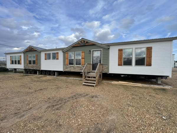 A large manufactured home with gray siding and brown shutters sits on a gravel lot under a cloudy sky. Wooden steps lead to the front door.