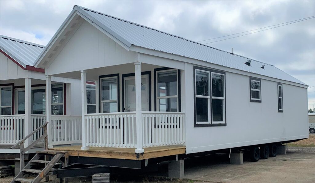 A small white manufactured house with a metal roof and front porch. It rests on a platform with stairs, surrounded by overcast sky, conveying simplicity.