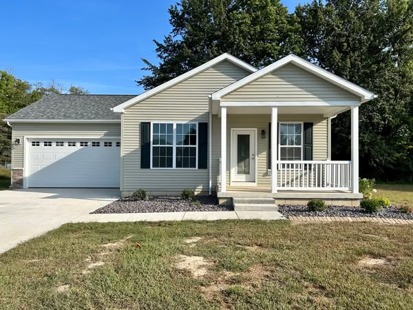 Single-story beige house with white trim, a small front porch, and a two-car garage. Well-kept lawn, a concrete driveway, and surrounded by tall trees.