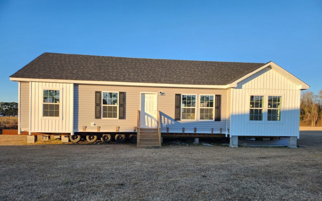 A beige modular home with black shutters sits elevated on a grassy field, illuminated by warm sunlight. A small staircase leads to its white front door.