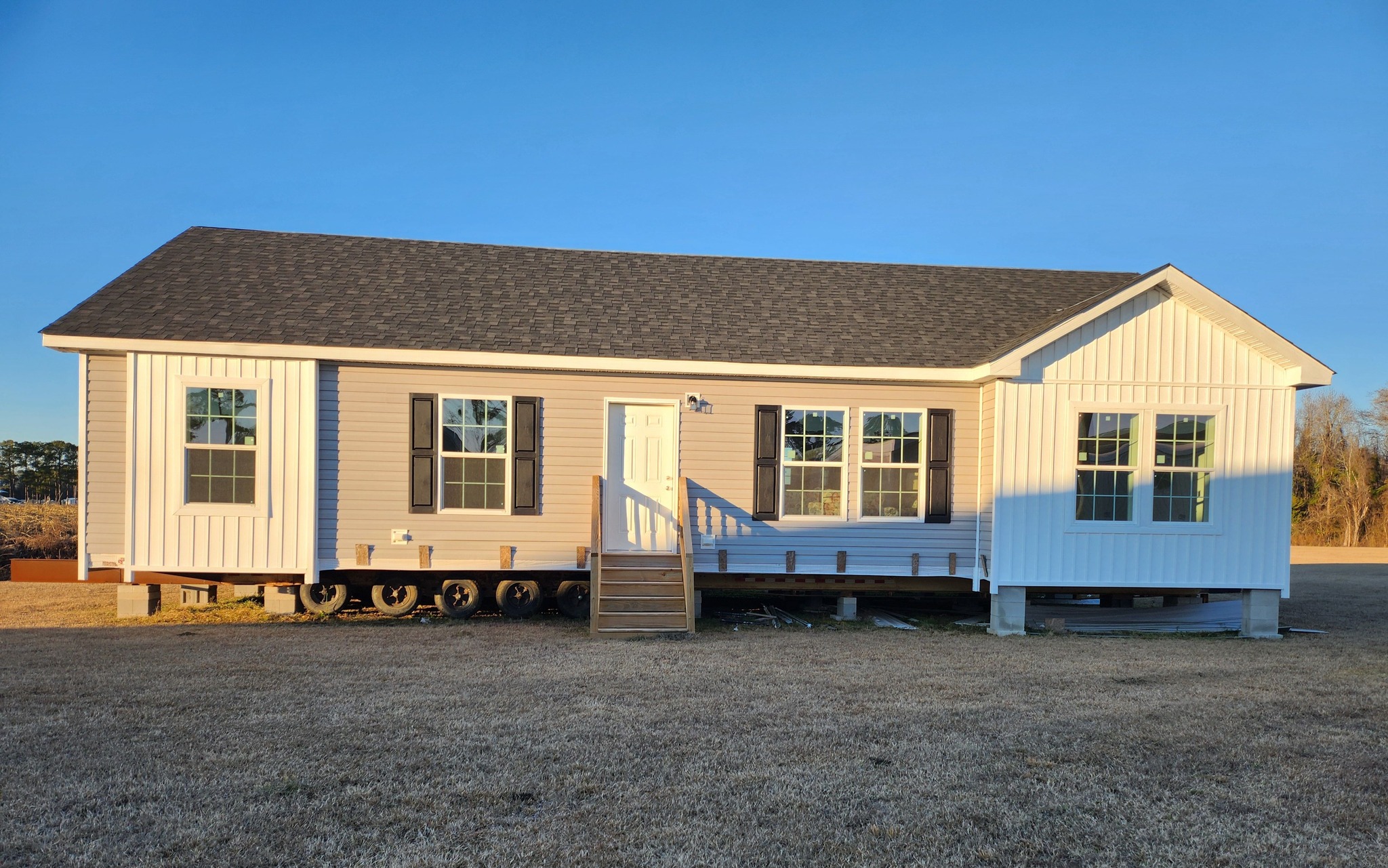 A beige modular home with black shutters sits elevated on a grassy field, illuminated by warm sunlight. A small staircase leads to its white front door.