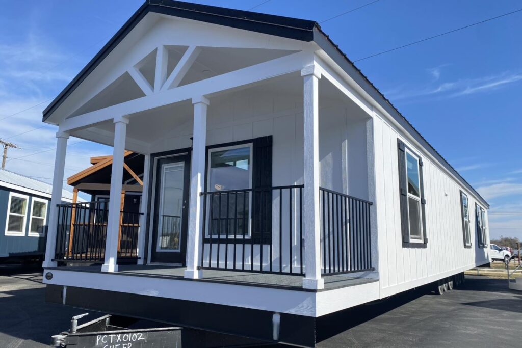 Modern white manufactured home with black trim, featuring a covered porch with railings. Set on a lot under a clear blue sky, evoking a sense of simplicity.