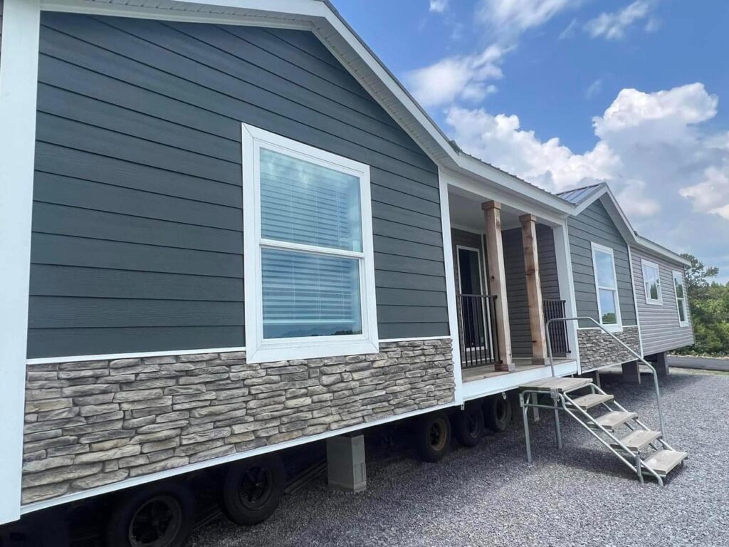 Single-story mobile home with dark gray siding and stone accents. It has large windows, a covered porch with wooden columns, and metal steps leading up. Sky is partly cloudy.
