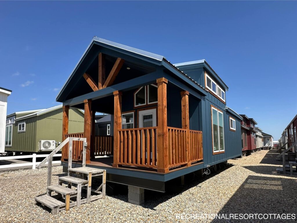Small blue cabin with a wooden porch, set on a gravel lot and lit by bright sunlight. Other cabins are visible in the background on a clear day.