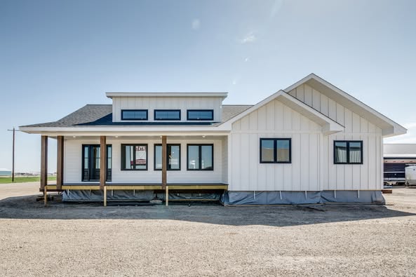 Single-story white house with modern design and dark-framed windows. The porch features wooden posts, set against a clear blue sky and gravel foreground.