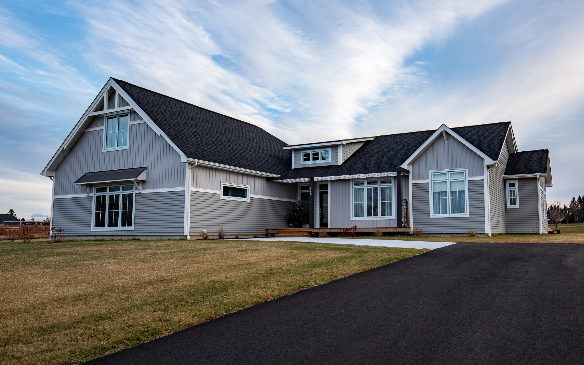 A modern single-story gray house with white trim sits under a blue sky. It features large windows, a dark roof, and a spacious driveway, conveying a calm suburban feel.