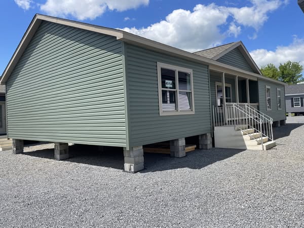 A green, single-story manufactured home on a gravel lot under a partly cloudy blue sky. The home is elevated on cinder blocks with a small white staircase leading to the entrance.