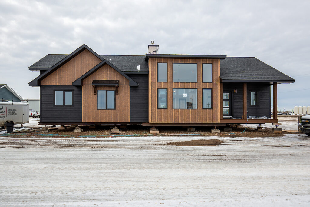 Modern black and wood-toned modular home on a snowy lot with large windows. The structure is elevated on blocks, showcasing an industrial setting.