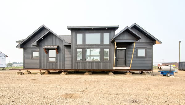 A modern, dark gray modular home sits elevated on blocks against a barren, dirt landscape. Large windows dominate the facade, suggesting a bright interior.